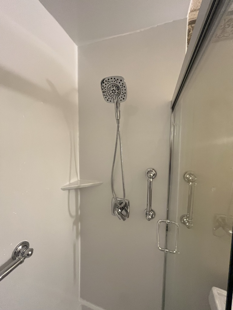 Shower detail with a handheld shower head on a slide bar, a corner shelf and two grab bars