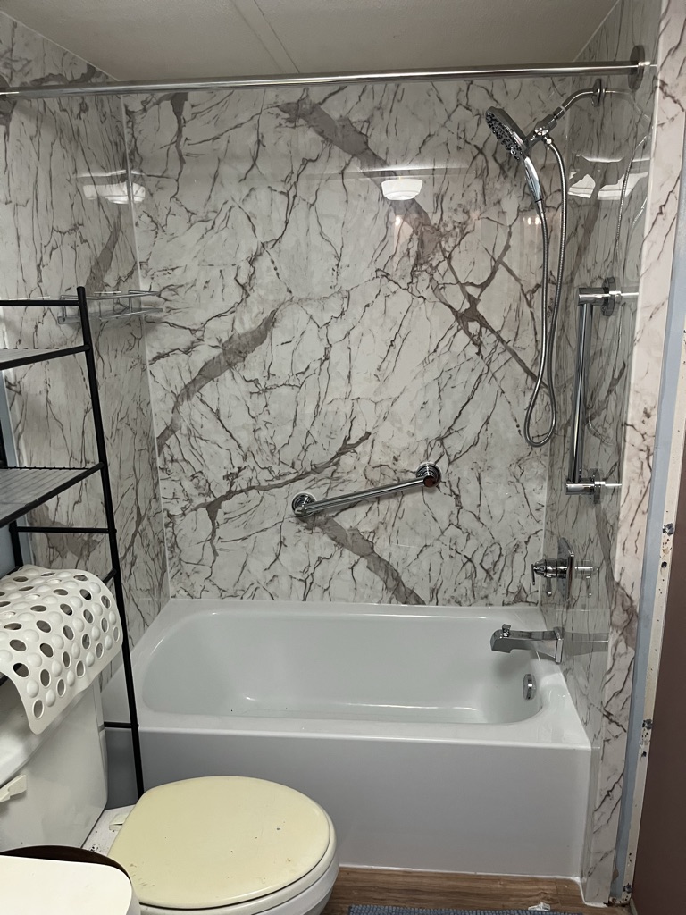 Tub and shower with marble look wall panels, a grab bar and a handheld shower on a slide bar