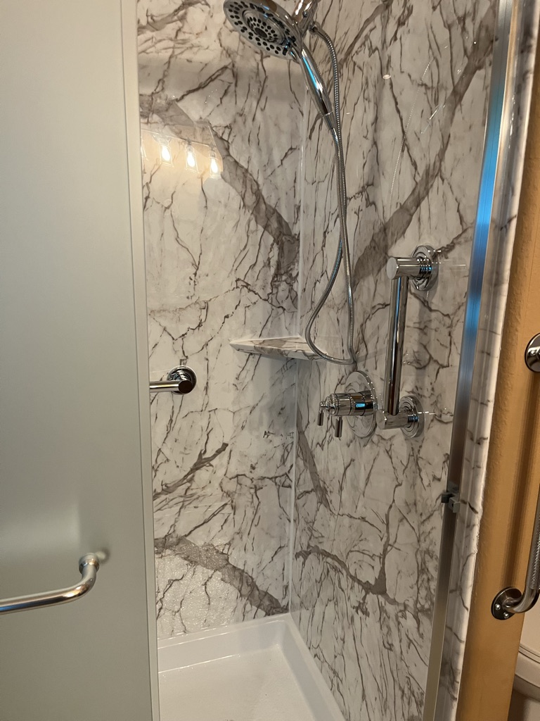 Shower detail with marble look walls, a handheld shower head, corner shelf and grab bars