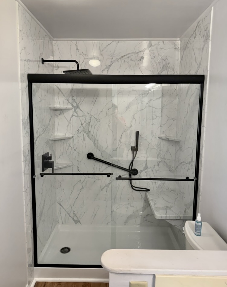 Walk in shower with marble look walls, a bench, corner shelves and a black framed sliding glass door