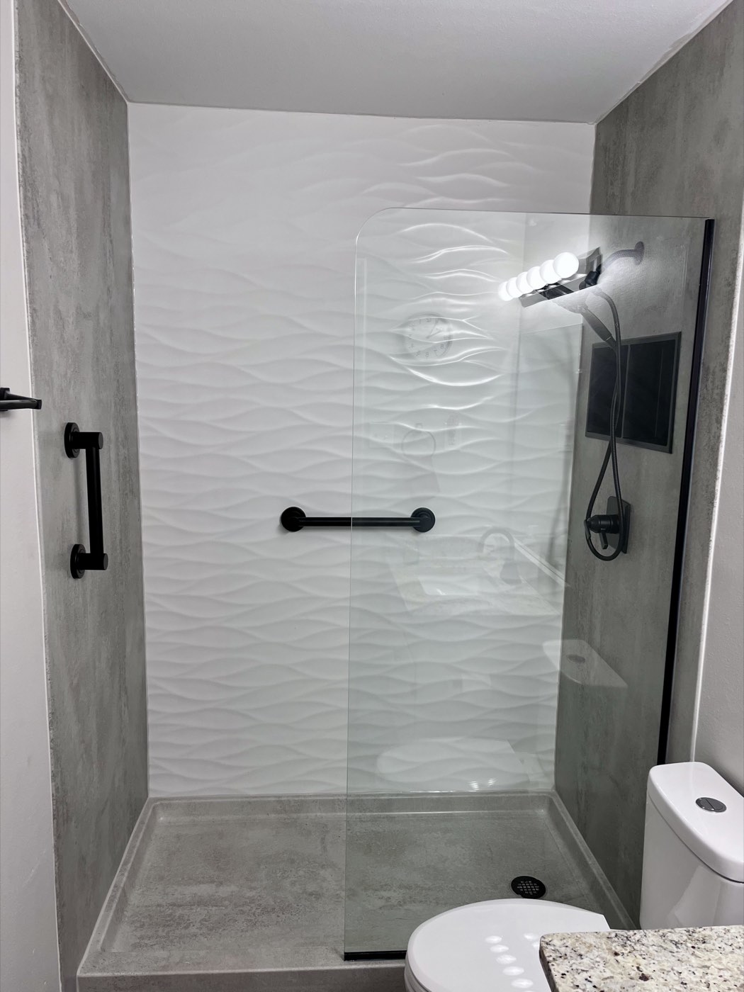 Walk in shower with a wave pattern back wall, concrete look side walls, a glass panel and black grab bars